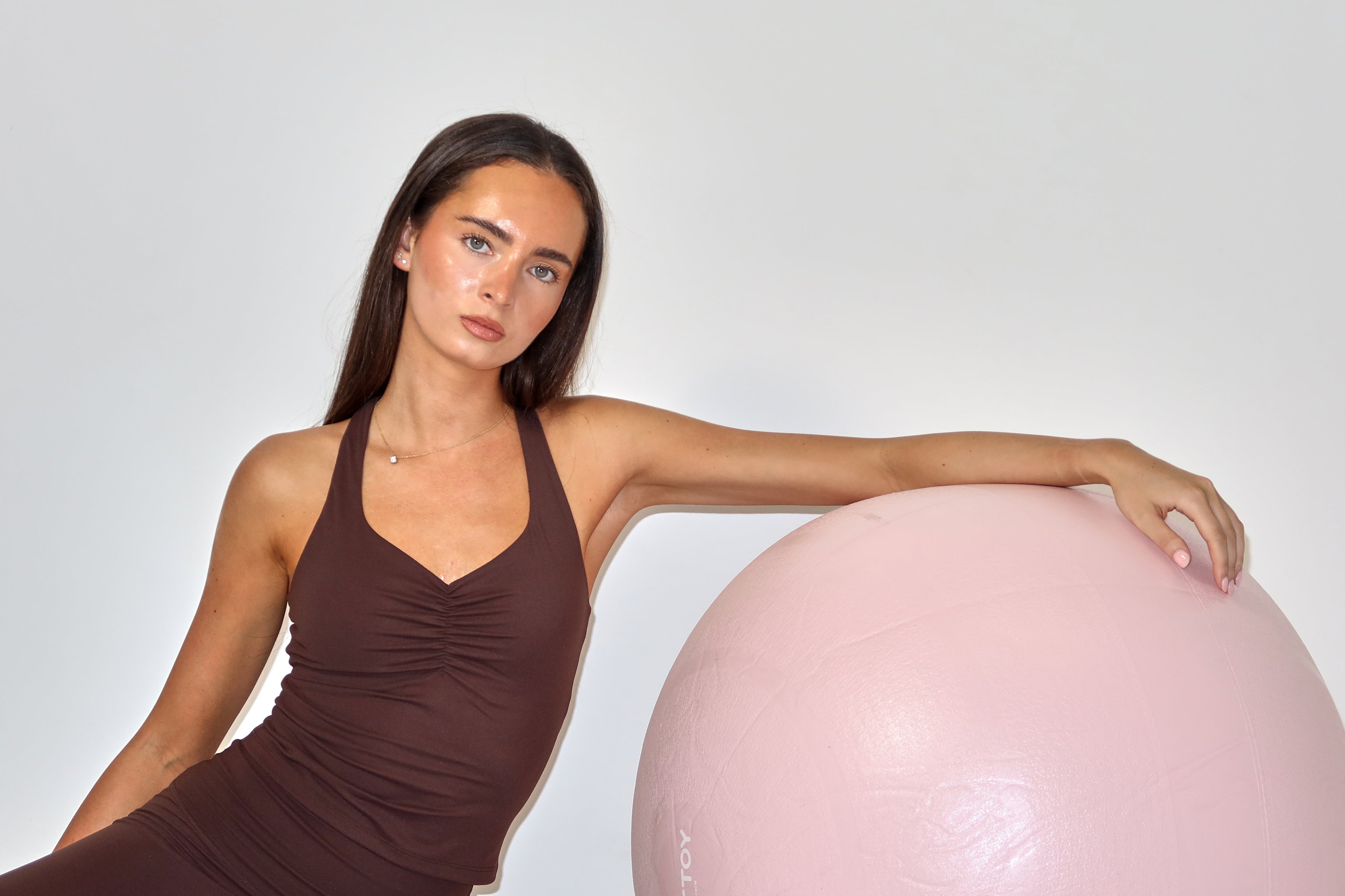 Woman in a brown dress leaning against a pink exercise ball on a white background in activewear raae uk